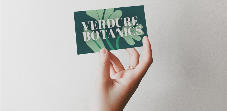 Ruka drží modernú vizitku s nápisom „Verdure Botanics“ s botanickým dizajnom na zelenom pozadí.