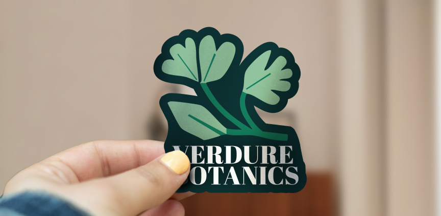 Ruka drží samolepku s logom „Verdure Botanics“ a ilustráciou listov v zelených odtieňoch na neutrálnom pozadí.