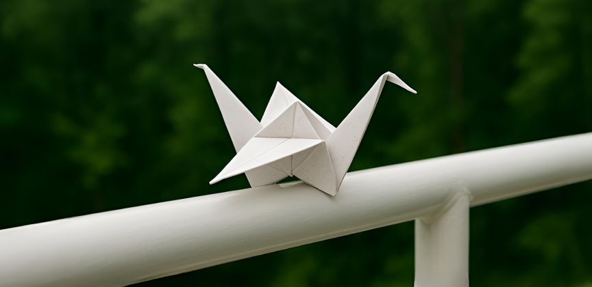Papierový origami žeriav položený na bielom zábradlí s prírodou v pozadí.